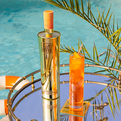 One Bottle of Au Vodka Juicy Peach, with a Au Highrise Glass filled with ice &  orange liquid. Placed on a reflective surface, next to an outdoor pool with rubber rings.