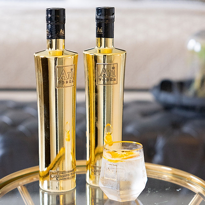 Two Bottles of Au Vodka Original on a reflective surface, with a diamond cut glass, full of clear liquid and a lemon slice garnish.