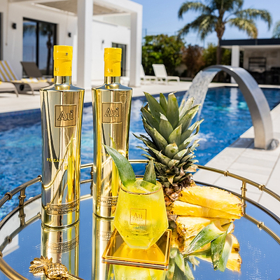 Two Bottles of Au Vodka Pineapple Crush, next to a Au Diamond Cut Glass, filled with Yellow Liquid. Next to a slice of pineapple garnish. On reflective surface next to an outdoor pool.