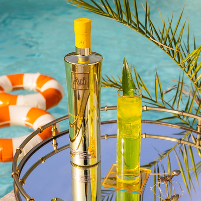 One Bottle of Au Vodka Pineapple Crush, with a Au Highrise Glass filled with ice & yellow liquid. Placed on a reflective surface, next to an outdoor pool with rubber rings. 
