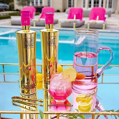 Two Bottles of Au Vodka Pink Lemonade, on a reflective surface, next to an outdoor pool. There's an Au Diamond Cut Glass, filled with ice, pink liquid and a lemon slice garnish. 