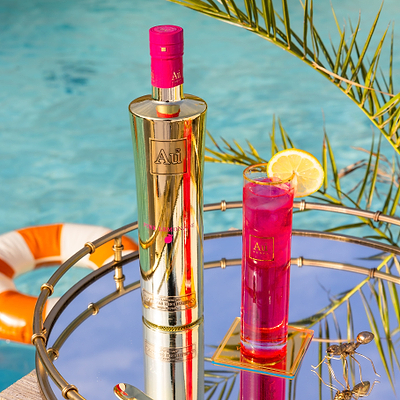 One Bottle of Au Vodka Pink Lemonade, with a Au Highrise Glass filled with ice & pink liquid. Placed on a reflective surface, next to an outdoor pool with rubber rings. 