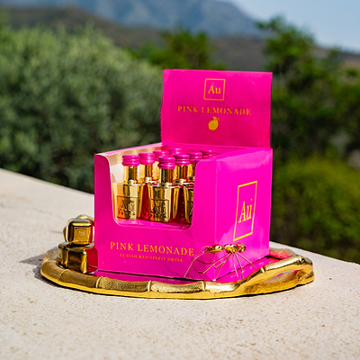12 Miniature Bottles of Au Vodka Pink Lemonade, in Pink Packaging, on a gold plate, in a outdoor summer setting.
