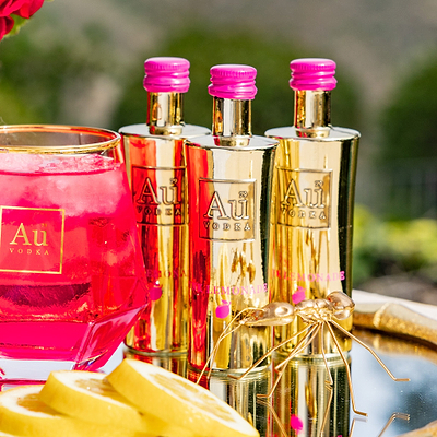 Three bottles of Au Vodka Pink Lemonade 50ml with a pink liquid and lemon slices on a reflective surface.