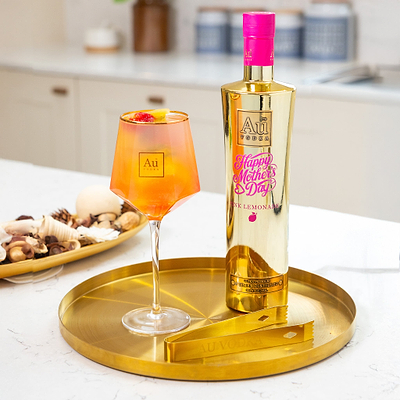 Bottle of Au Vodka Pink Lemonade, with a UV Print Design saying 'Happy Mother's Day' in Pink. Next to a long stem cocktail glass, on a gold serving dish, in a white marble kitchen.