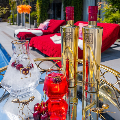 Two Bottles of Au Vodka Red Cherry, an Au Branded Glass of Red Liquid, a Decanter of ice, in an outdoor poolside setting.