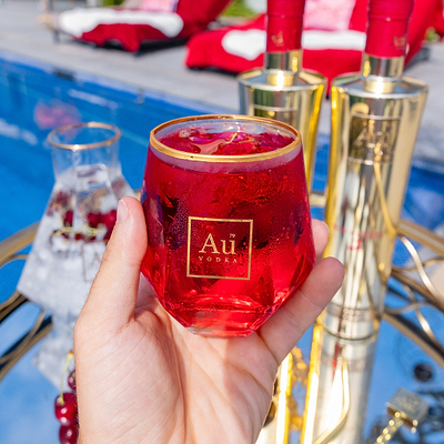 Hand Holding a Au Vodka Diamond Cut Glass, filled with Au Vodka Red Cherry, with a summery poolside background.