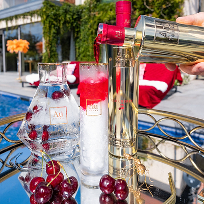 A bottle of Au Vodka Red Cherry being poured into an Au Highrise Glass full of ice, next to an outdoor pool in Summer.