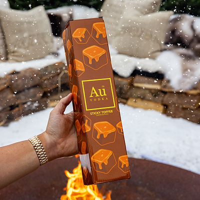 Hand holding Au Vodka Sticky Toffee Collection Box by a fire with snow in the background