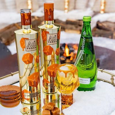 Two Bottles of Au Vodka Sticky Toffee with Limited Edition Toffee Glass. Next to a Bottle of Appletiser on a reflective surface. 