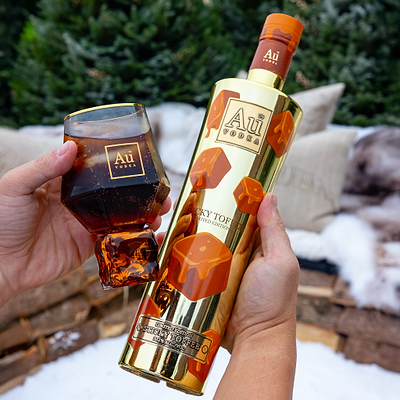Person holding a bottle of Au Vodka Limited Edition Sticky Toffee, and a glass of Au Vodka. 