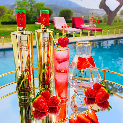 Two Bottles of Strawberry Burst next to a Highrise Glass filled with Red Liquid, and fresh strawberries on a reflective surface, next to an outdoor pool.