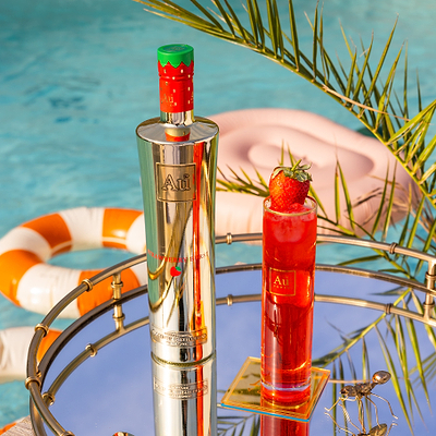 One Bottle of Au Vodka Strawberry Burst, with a Au Highrise Glass filled with ice & red liquid. Placed on a reflective surface, next to an outdoor pool with rubber rings. 