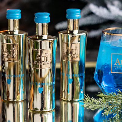 Four Au Vodka Blue Raspberry Miniature 50ml Bottles, next to an Au branded glass of blue liquid, on a reflective surface. 