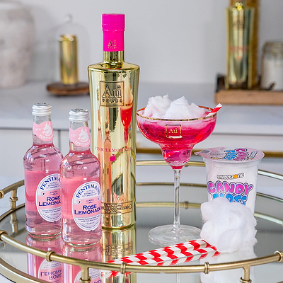 Bottle of Aul Gold Liqueur with pink cap, two bottles of Pink Lemonade, a pink cocktail in a glass with ice, and a container of candy on a gold tray.