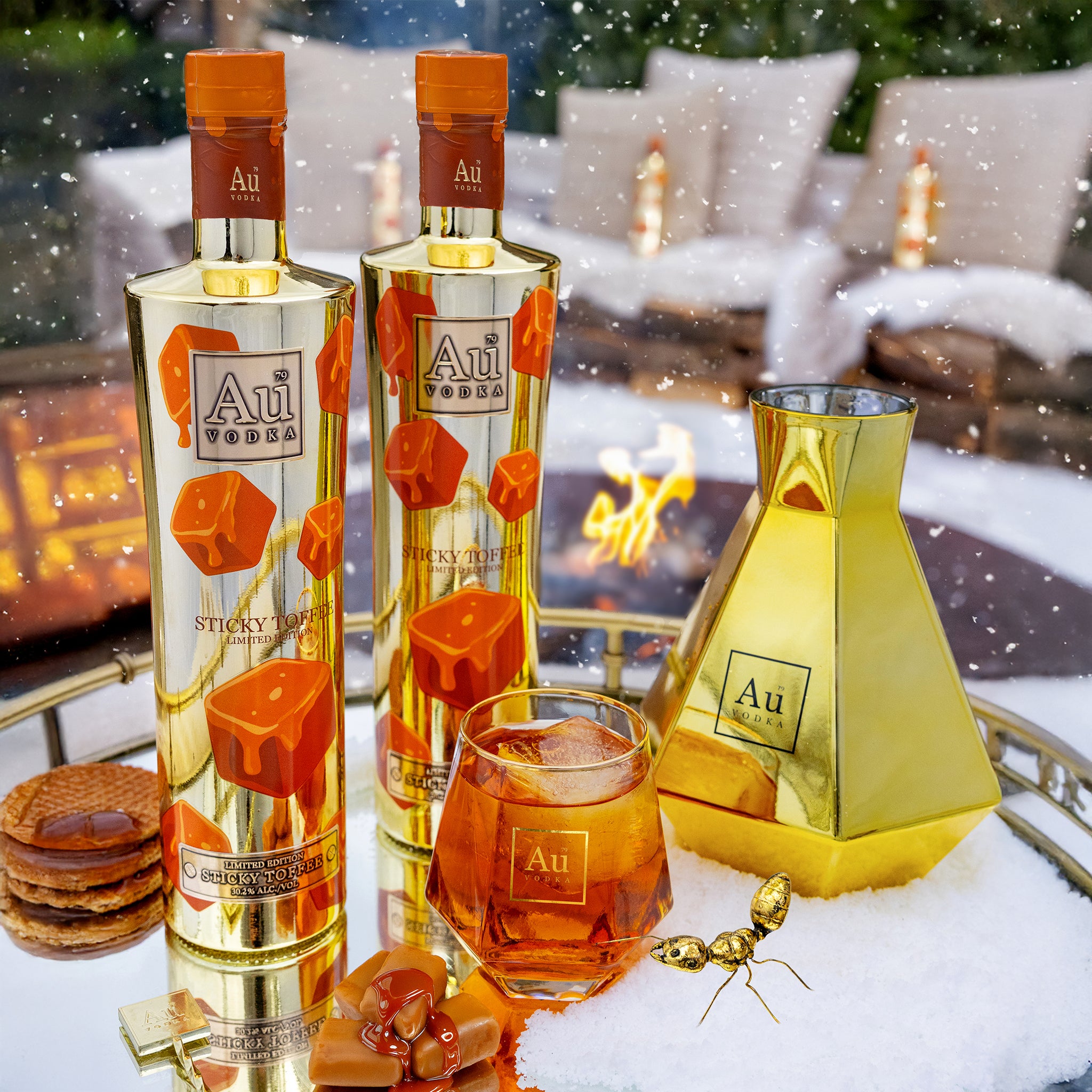 Two bottles of Au Vodka Sticky Toffee with a glass of the drink in front of a snowy outdoor setting with a fire pit.
