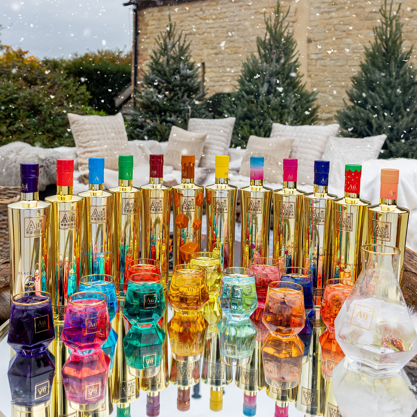 Colorful bottles with gold accents on a reflective surface outdoors with trees and a building in the background.