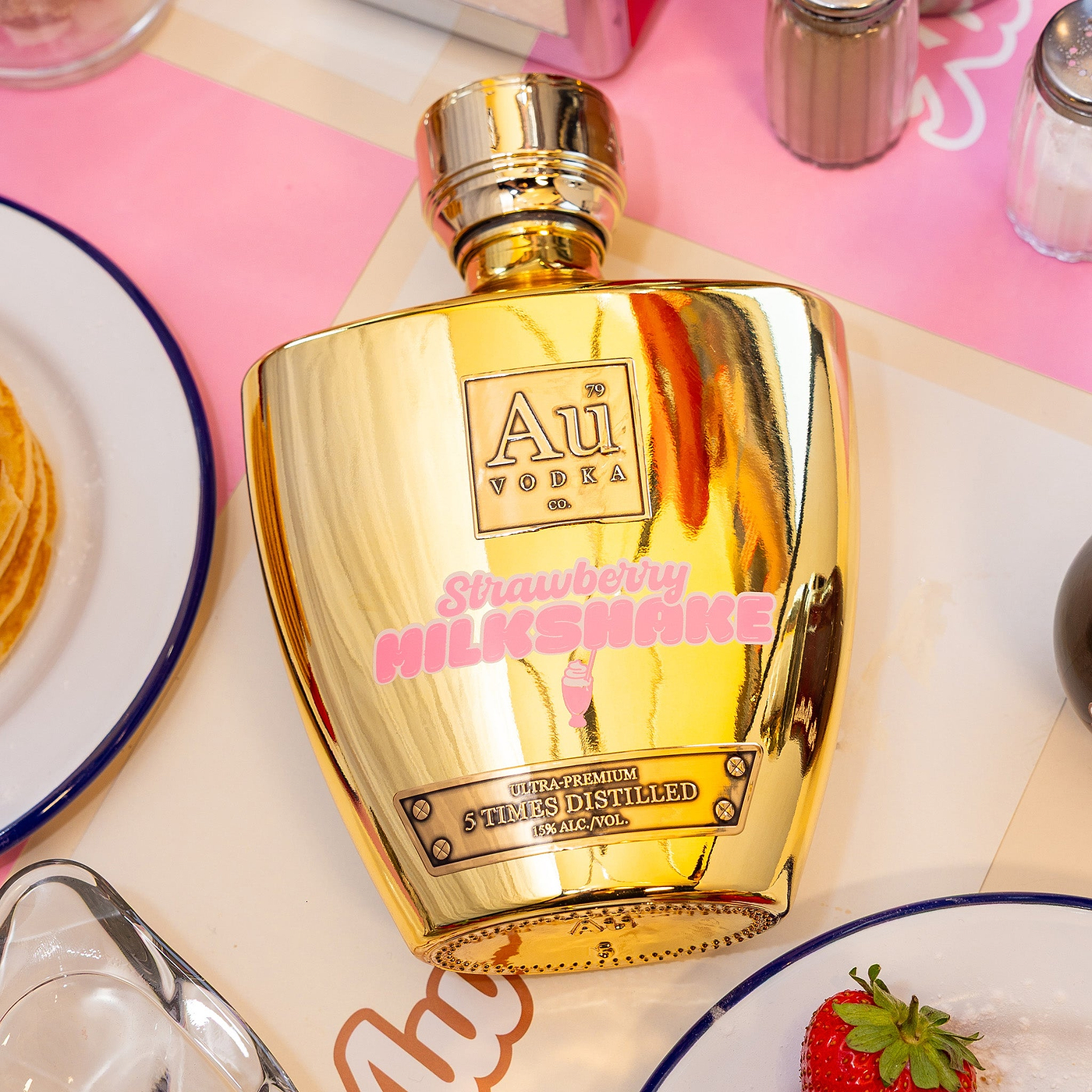Gold bottle of Au Vodka Strawberry Milkshake flavour on a table with plates and a strawberry.