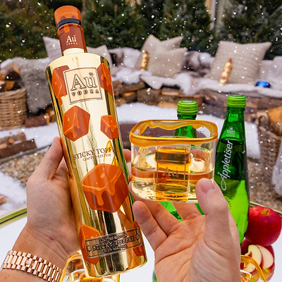 Person holding a bottle of Au Vodka Sticky Toffee and a glass with Appletiser, outdoor setting with Christmas decorations.