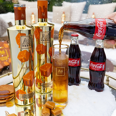 Two bottles of Au Vodka being poured into a glass with Coca-Cola bottles in the background.