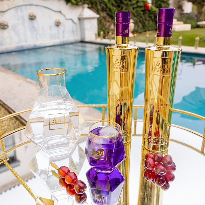 Two Bottles of Au Vodka Black Grape, next to a clear decanter & diamond cut glass with purple liquid. Set on a reflective surface, next to an outdoor pool backdrop. 