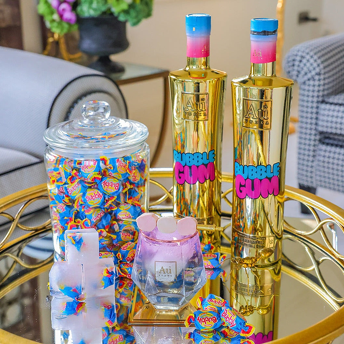 Au Vodka Bubblegum on a reflective surface, with a diamond cut glass, filled with bubblegum liquid and a sweet garnish, on a reflective surface.