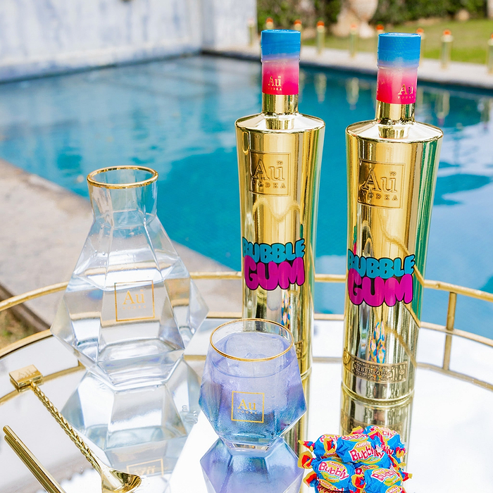 Two bottles of Au Vodka Bubblegum displayed on a reflective tray with a clear glass and bubblegum flavored candy, with a pool in the background