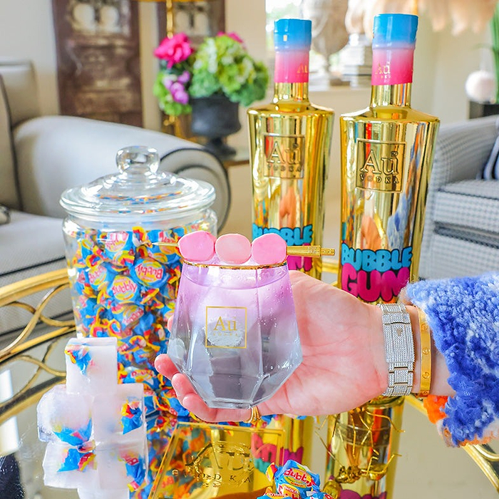 A hand holding a Diamond Cut Glass filled with Au Vodka Bubblegum, with a sweet garnish, with a sweet jar and two bottles of Au Vodka Bubblegum in the background. 