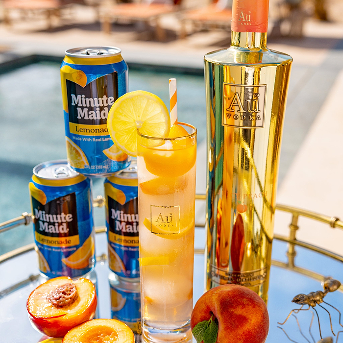 One bottle of Au Vodka Juicy Peach 750ml & an Au branded Glass full of oragne liquid and peach slices. Next to three cans of Minute Maid, on a reflective surface. 