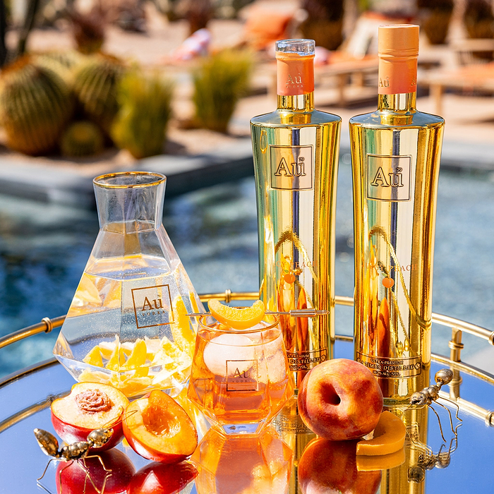 Two bottles of Au Vodka Juicy Peach next to an Au Vodka Diamond caut Glass, filled with orange liquid and peach slices, on a reflective surface. 