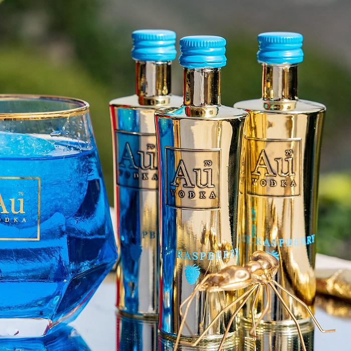Three miniature bottles of Au Vodka Blue Raspberry in gold-colored bottles with blue caps, displayed alongside a glass of blue liquid. 
