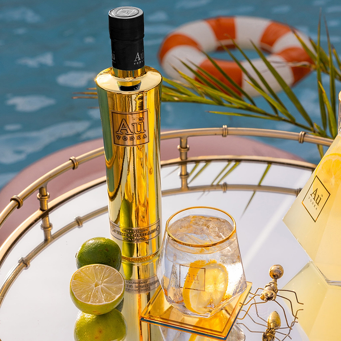 A Bottle of Au Vodka Original in a 375ml Bottle, next to an Au Diamond Cut Glass filled with vodka, and a slice of lemon, next to two halfs of a lime, on a reflective surface. 