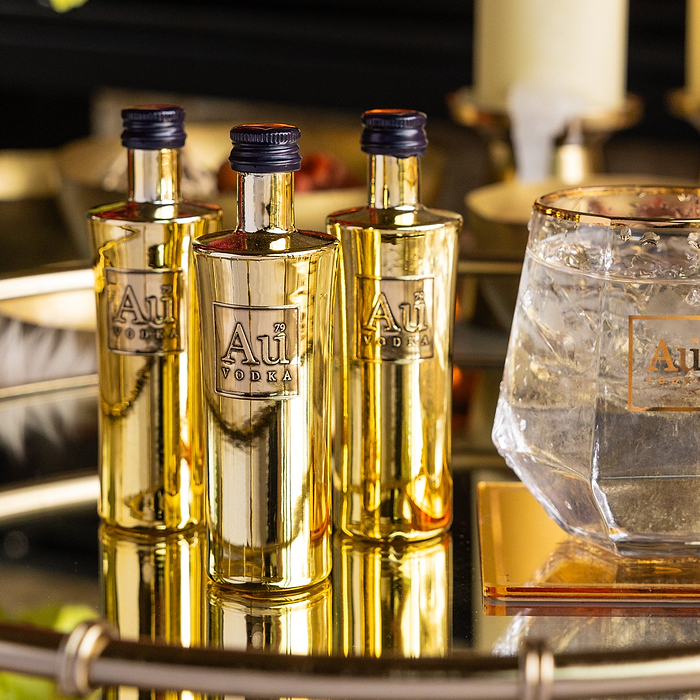 Three Miniature gold bottles of Au Vodka Original on a reflective surface with a glass containing ice and liquid.