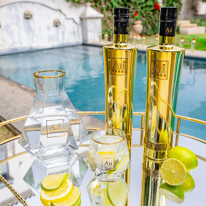 A Bottle of Au Vodka Original placed on a reflective surface with a glass decanter 7 diamond cut glass, lime slices, and ice cubes, set against a poolside backdrop.