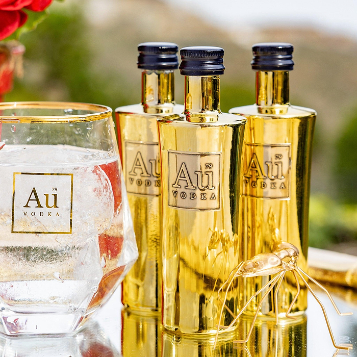 Three miniature bottles of Au Vodka Original placed behind a glass of ice water, all three bottles are sealed with black caps and have the brand name 'Au Vodka' displayed on them, with a gold-colored finish.