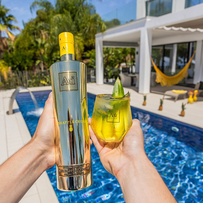 A Person holding a bottle of Au Vodka Pineapple Crush & Au Diamond Cut Glass, with yellow liquid, on an outdoor pool backdrop. 