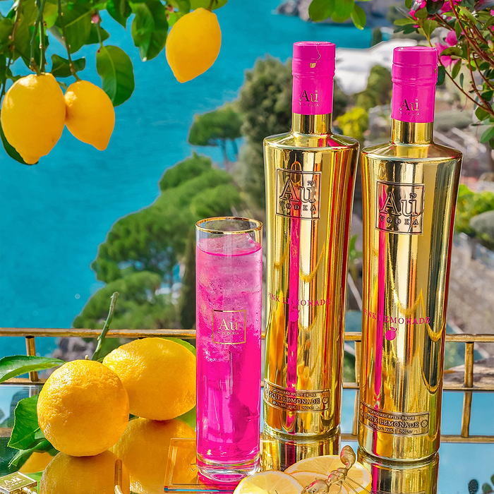 Two bottles of Au Vodka Pink Lemonade, with a Au Highrise Glass filled with Ice & Pink Liquid, placed on a reflective surface. With a scenic, outdoor backdrop