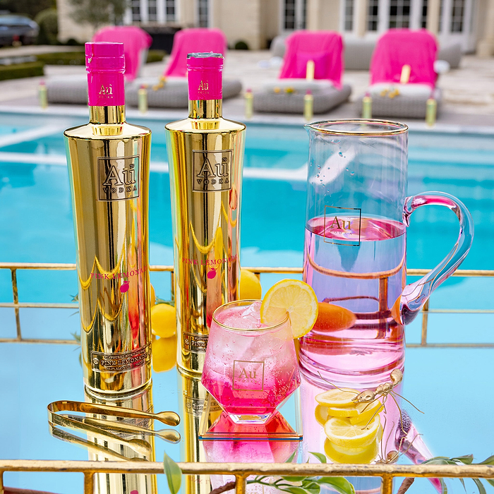 Two Bottles of Au Vodka Pink Lemonade, on a reflective surface, next to an outdoor pool. There's an Au Diamond Cut Glass, filled with ice, pink liquid and a lemon slice garnish. 