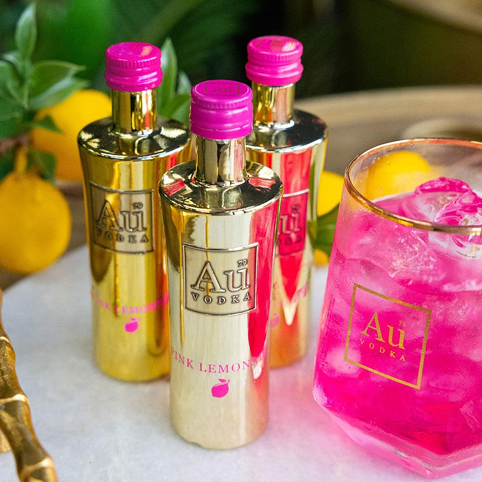 Three bottles of Au Vodka Pink Lemonade 50ml Miniature & glass filled with pink liquid on a marble surface.