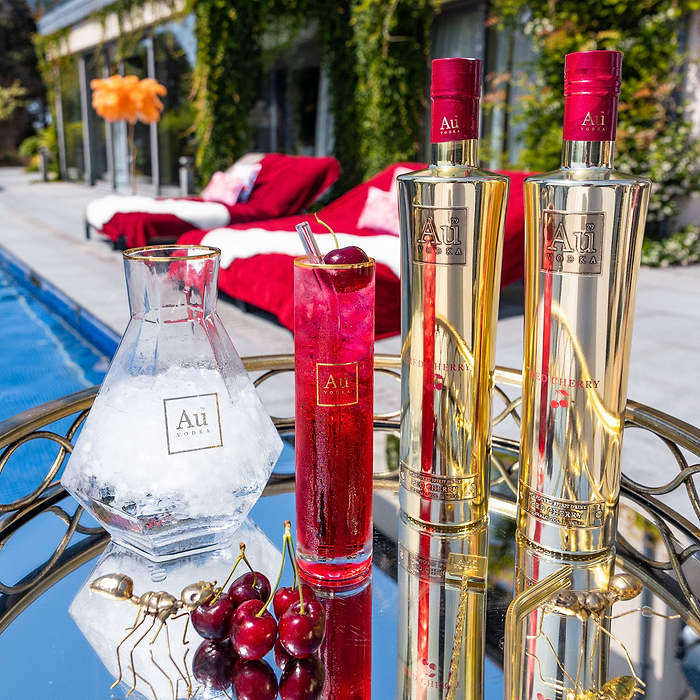 Two Bottles of Au Vodka Red Cherry, next to Au Highrise Glass, full of red liquid. Beside an outdoor pool in the summer time.