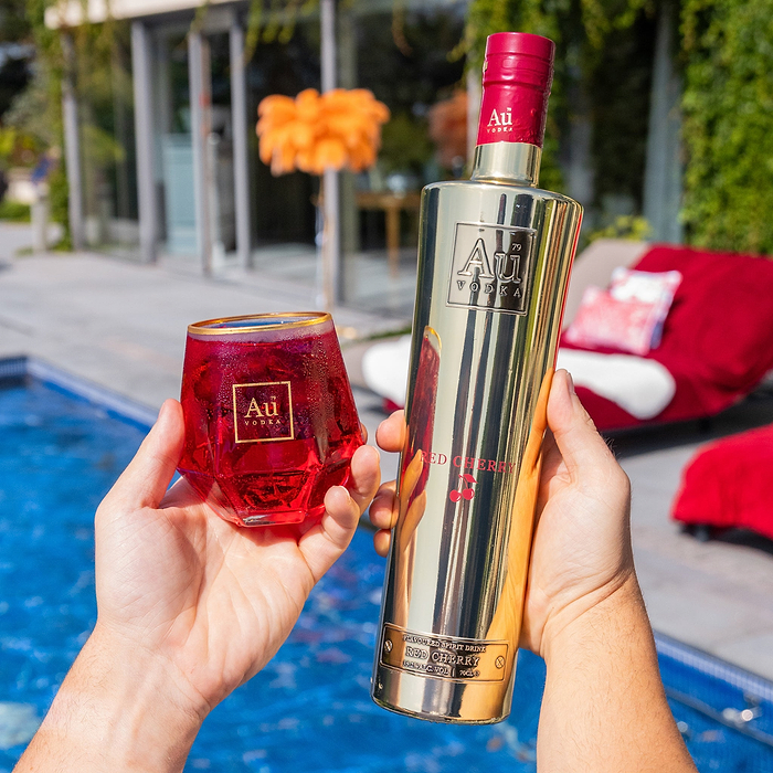 A hand holding a Bottle of Au Vodka Red Cherry & a Au Branded Diamond Cut Glass. Beside a Outdoor pool in summer time.