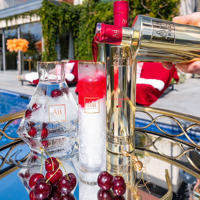 A bottle of Au Vodka Red Cherry being poured into an Au Highrise Glass full of ice, next to an outdoor pool in Summer.