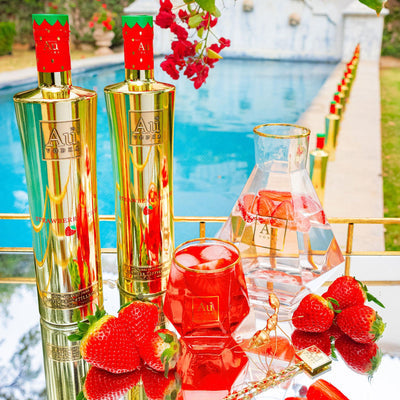 Two Bottles of Au Vodka Strawberry Burst, Diamond Cut Glass with red liquid. On a reflective surface, next to an outdoor pool