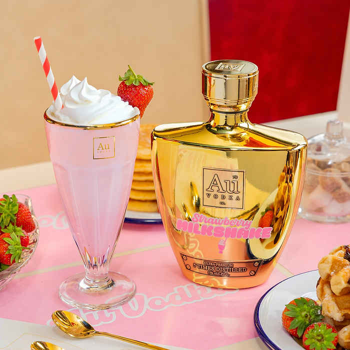 Au Vodka Strawberry Milkshake Bottle, next to a Au Sundae Glass, with whipped cream & a strawberry, on a pink diners table with dessert items.
