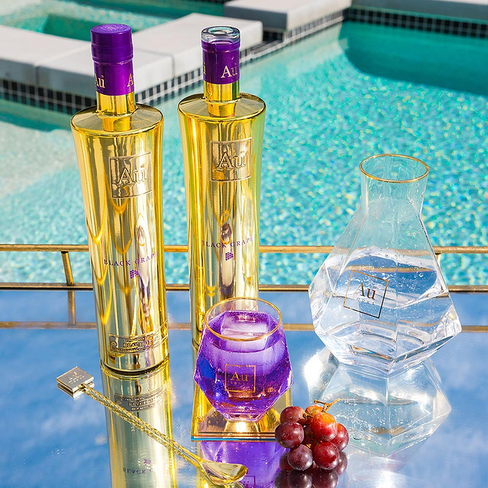 Au Vodka Black Grape 750ml on a poolside backdrop, with a Au Diamond Cut glass, full of purple liquid and black grape garnish on a reflective surface. 