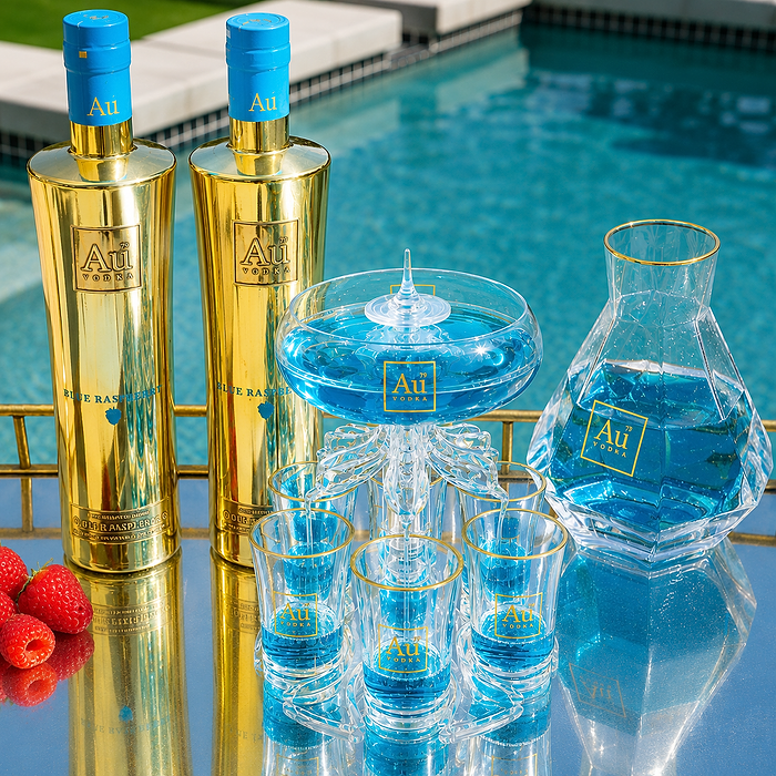 A luxury cocktail party set by Au Fine Spirits, featuring gold bottles of Raspberry Ketones, a glass pitcher and carafe filled with blue liquid, and matching gold-rimmed glasses and shot glasses, arranged on a reflective table next to raspberries with a swimming pool in the background.