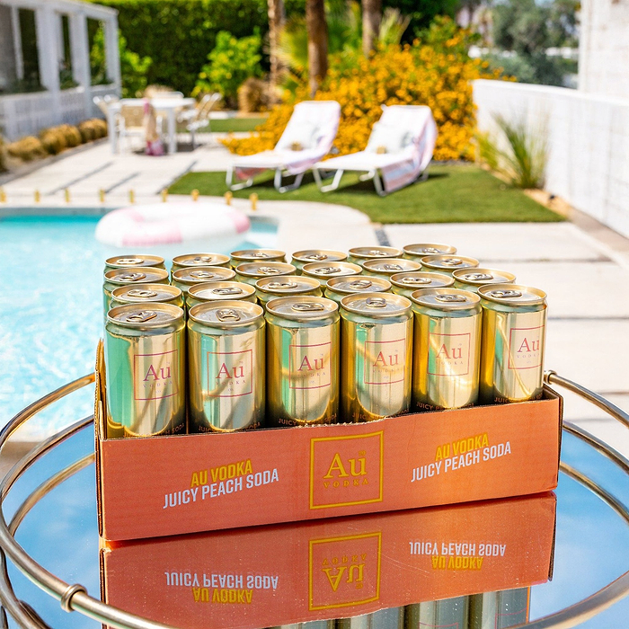 A 24-pack of Au Vodka USA Juicy Peach cans on a mirrored tray by a pool