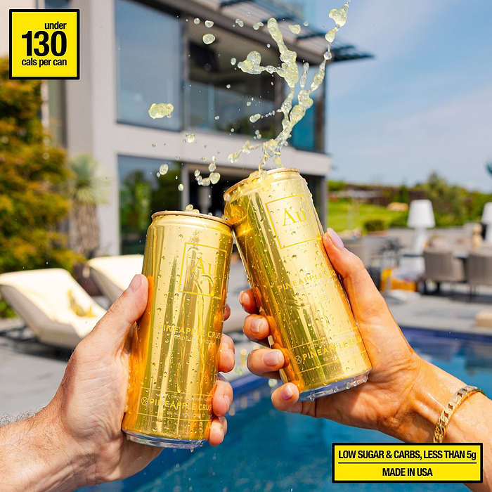 Two hands clinking Au Vodka USA Pineapple Crush cans with liquid splashing out, by a pool
