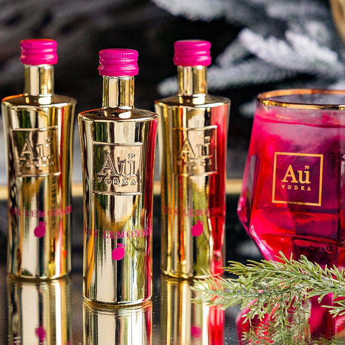 Three Miniature 50ml Au Vodka Bottles, Pink Lemonade. Next to a Au Vodka Glass of Pink Liquid on a glass surface.