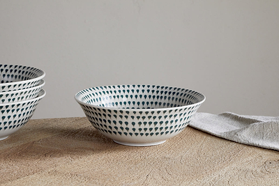 A white ceramic Juniper Drop Cereal Bowl in cream and juniper by nkuku, featuring a blue dotted pattern, sits on a wooden surface beside a folded beige cloth; more matching bowls are partially visible to the left—perfect for everyday dining.